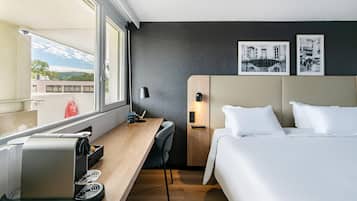 A hotel room with a bed, a desk, a chair, a lamp, and a wall-mounted control panel.