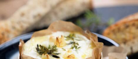 A dish of baked cheese with herbs, served in a paper-lined bowl, accompanied by bread on a dark plate.