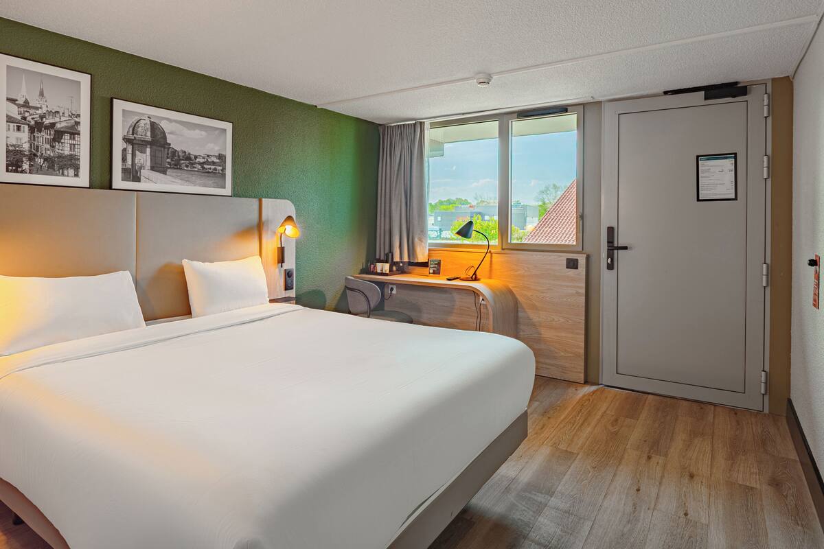 Een hotelkamer met een bed, een bureau, een stoel, een raam met uitzicht en twee ingelijste foto's aan de muur.