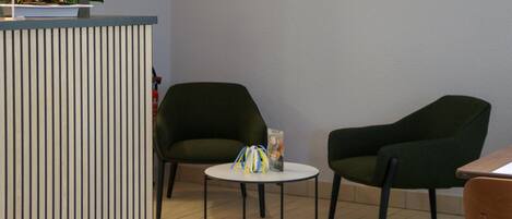 A modern interior with a reception desk, two green armchairs, a small round table, and a potted plant.