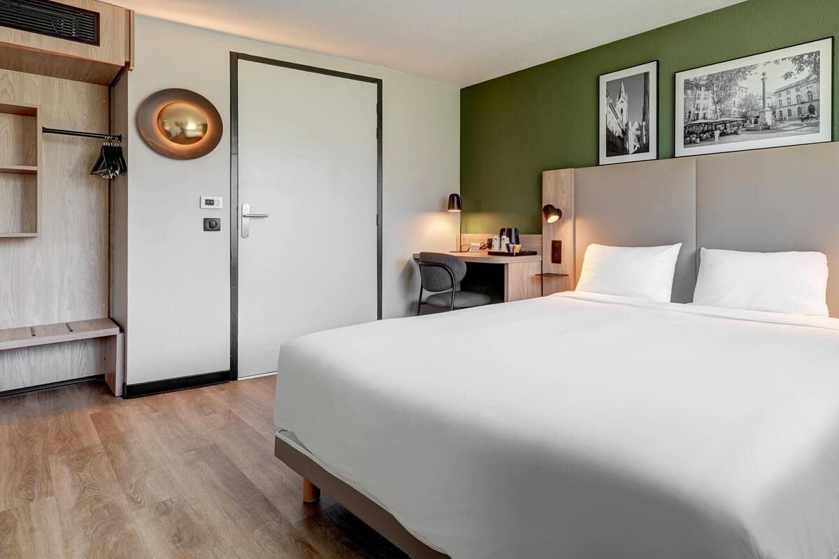 A hotel room with a large bed, a desk with a chair, a wardrobe, and two framed pictures on the wall.