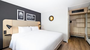 A modern hotel room with a large bed, a wooden headboard, a bedside table with a lamp, and a built-in wardrobe with shelves.