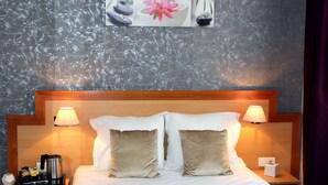 A neatly made bed with white and beige pillows, two bedside lamps, a coffee maker, and a small table with a box on it.