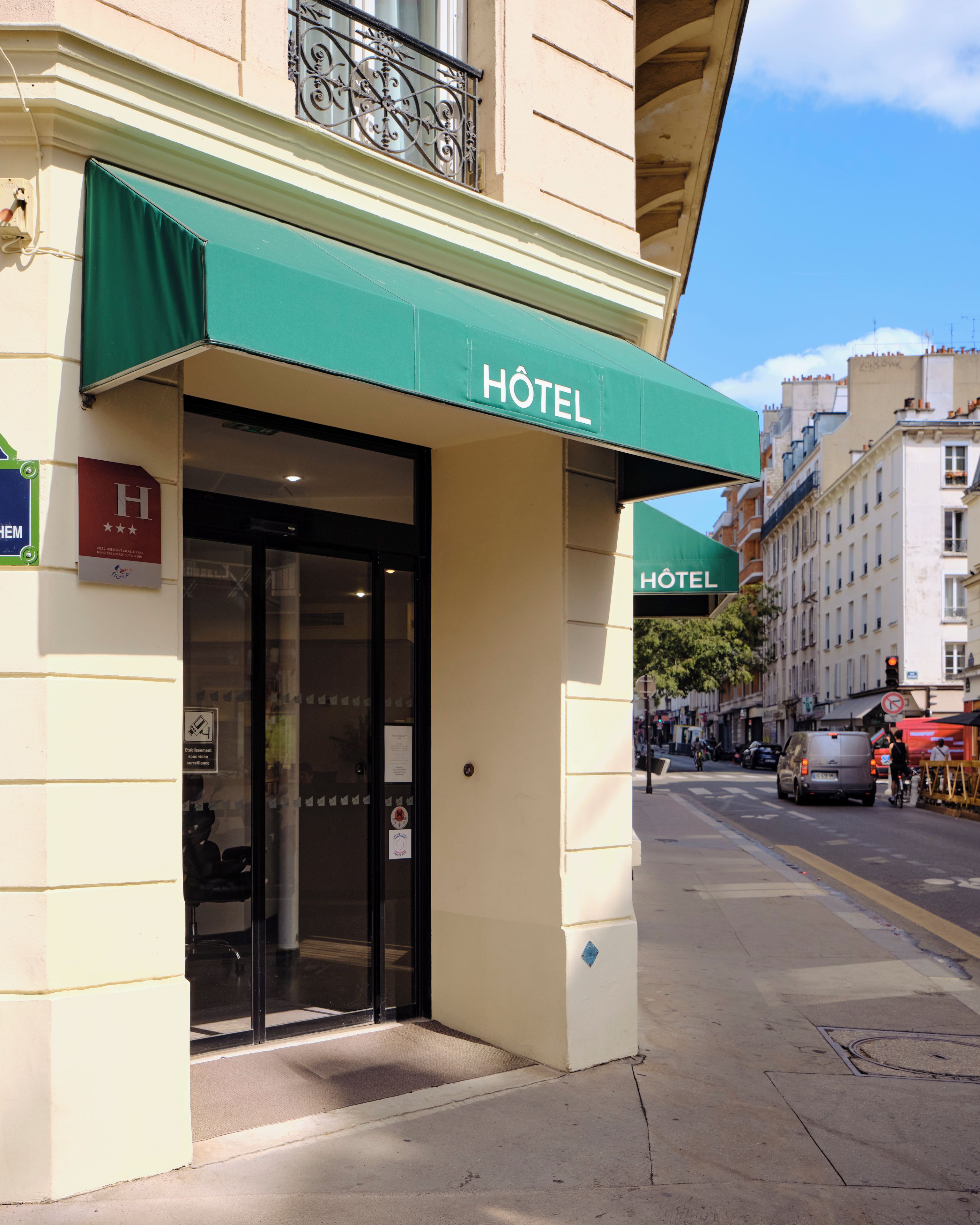 La façade d’un hôtel, avec un auvent vert, une entrée en verre et une vue sur la rue, où l’on aperçoit d’autres bâtiments et des véhicules.