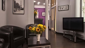 A reception area with a black leather sofa, a vase with yellow flowers, a flat-screen TV, and framed pictures on the walls.
