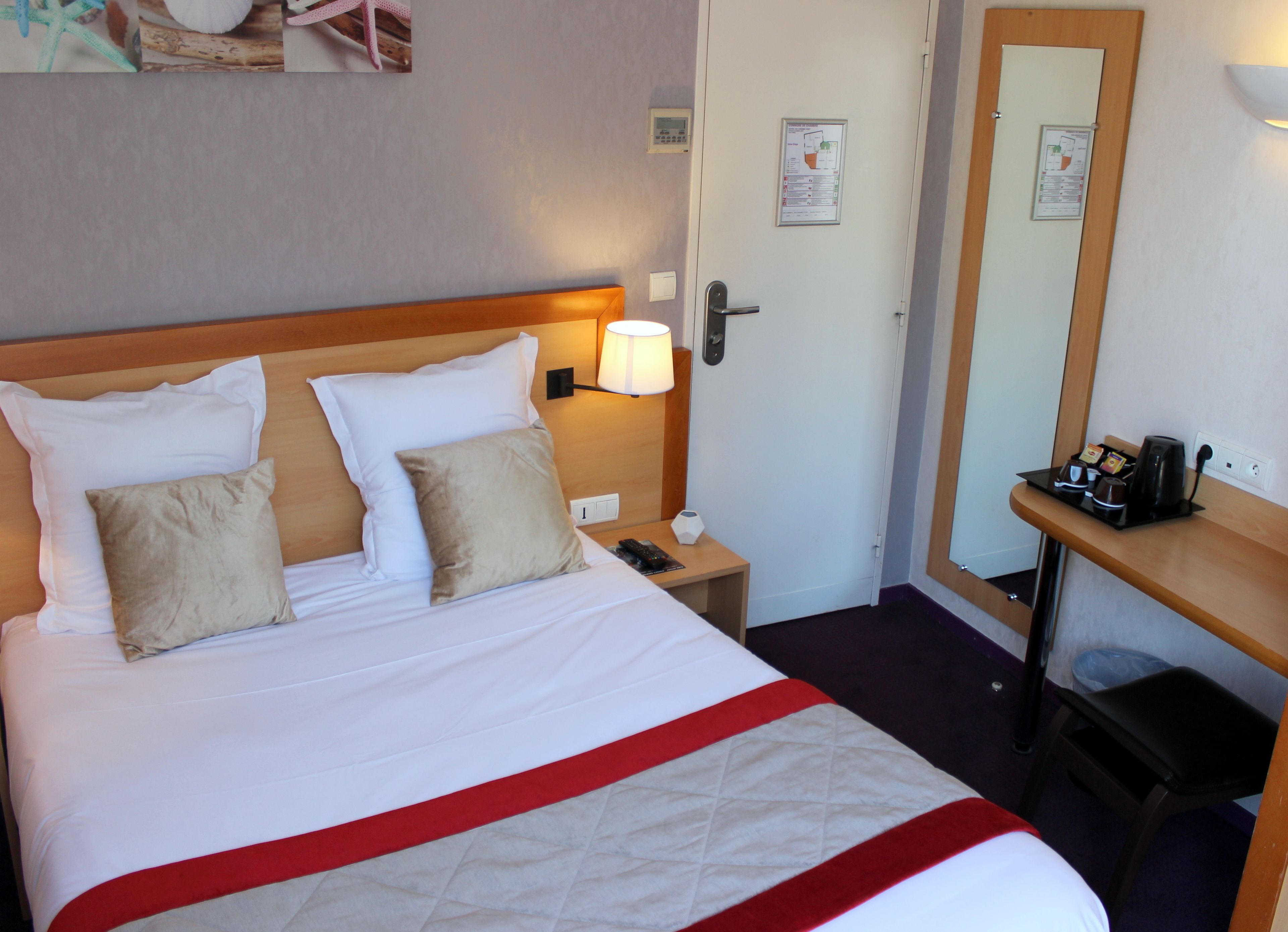 A hotel room with a bed, nightstand, lamp, mirror, and a small table with a coffee maker.
