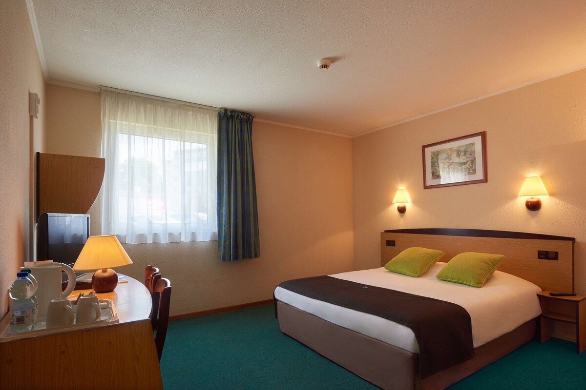 A hotel room with a bed, bedside lamps, a desk with a lamp, a coffee maker, and a window with curtains.