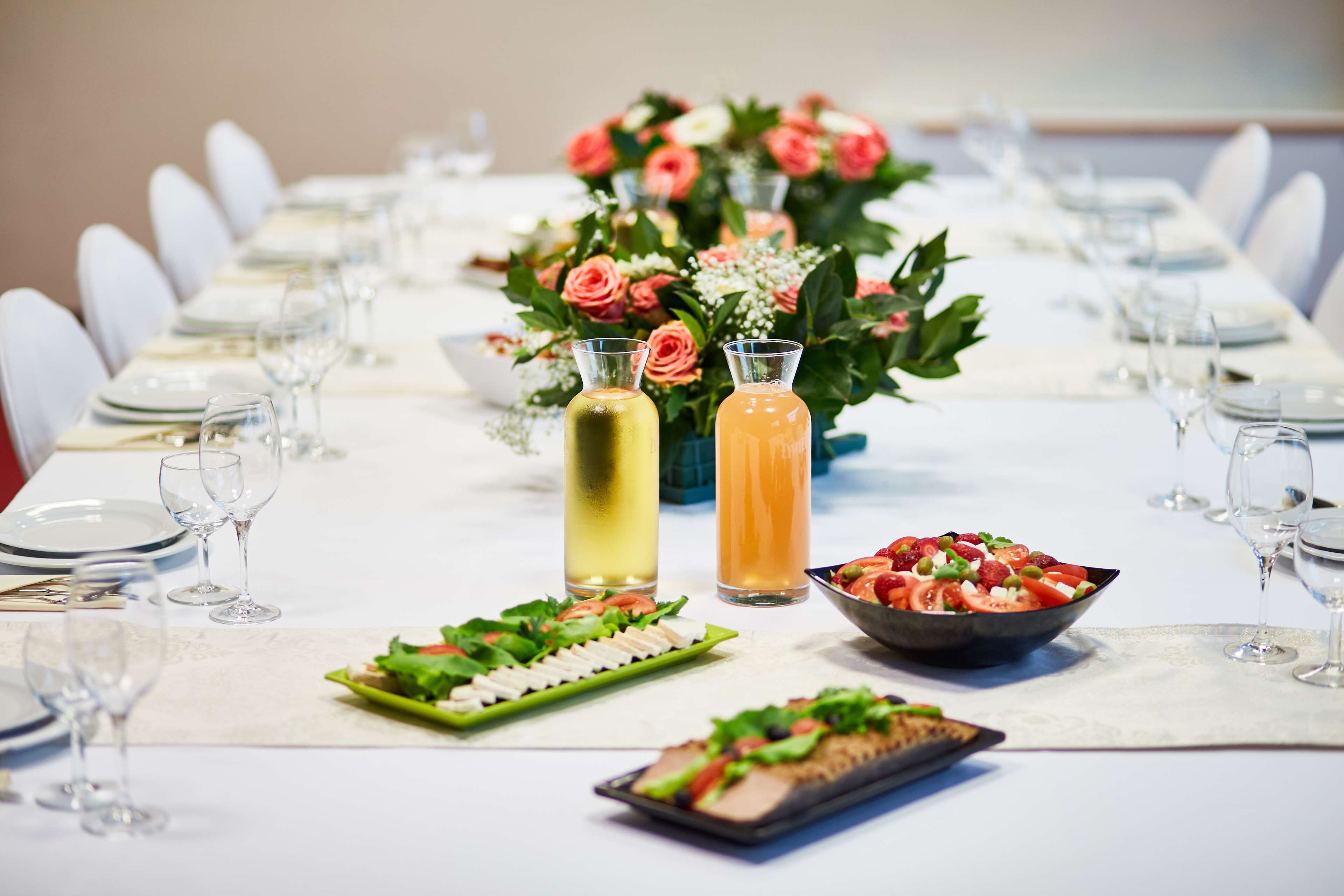 A long dining table set with white chairs, plates, and cutlery. There are two bottles of beverages and a bowl of salad.