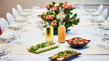 A long dining table set with white chairs, plates, and cutlery. There are two bottles of beverages and a bowl of salad.