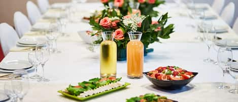 Ein langer Esstisch ist mit weißen Stühlen, Tellern und Besteck gedeckt. Zwei Flaschen Getränke und eine Salatschüssel stehen darauf.