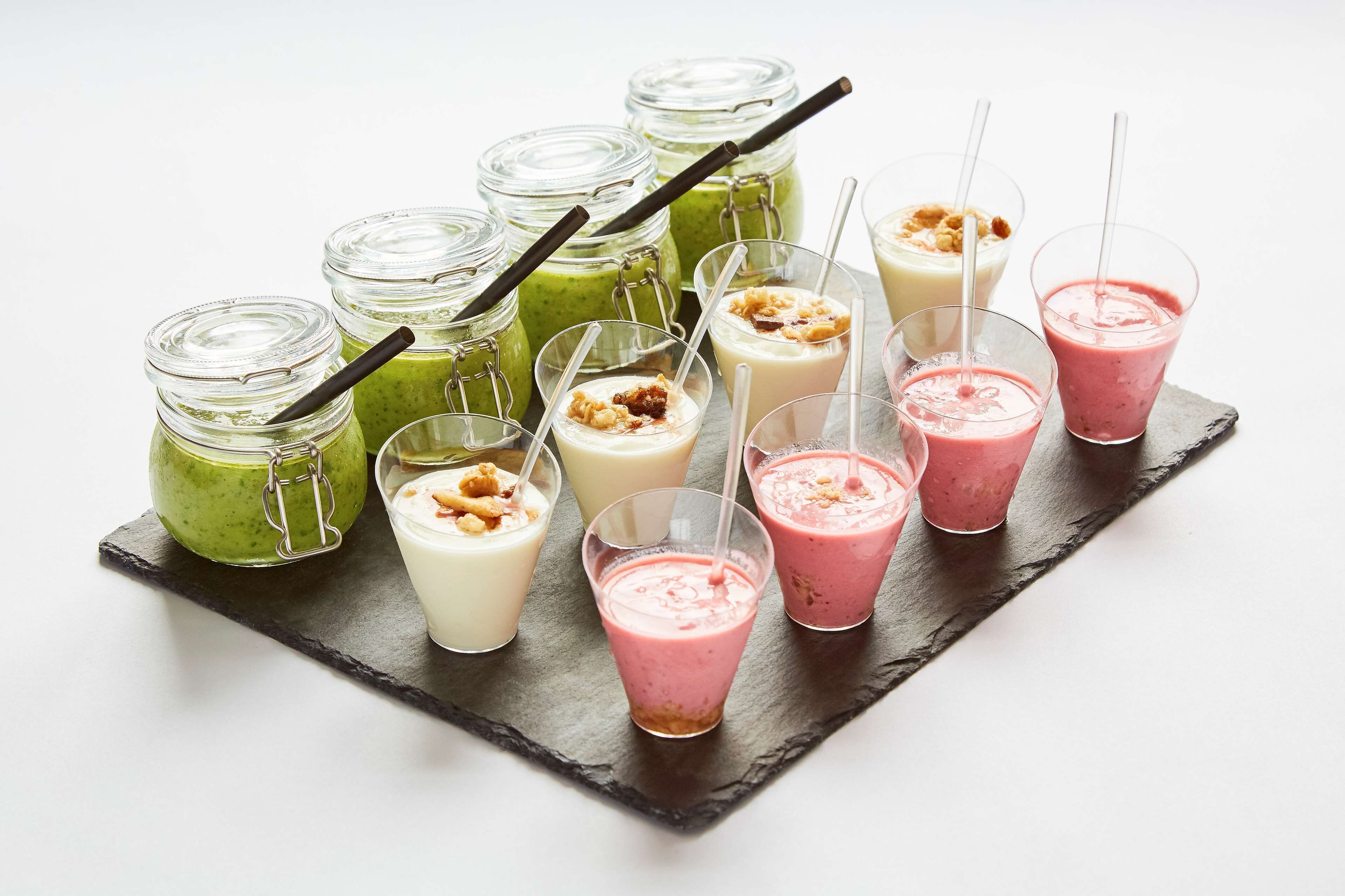 A variety of smoothies in different containers, including mason jars and glasses, on a slate board.