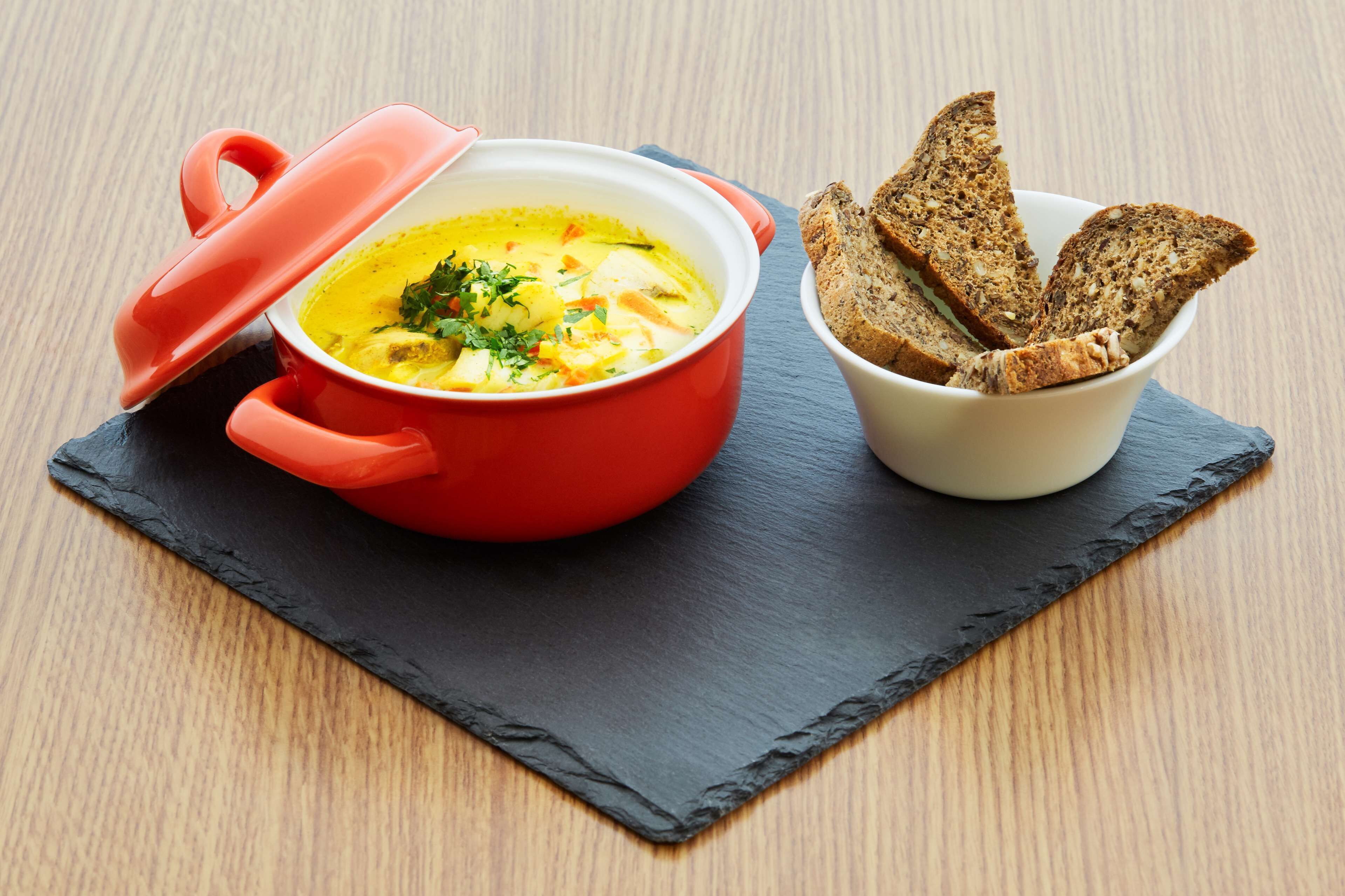 Een rode pan met deksel op een zwarte leisteenplaat, naast een kom met plakjes brood.
