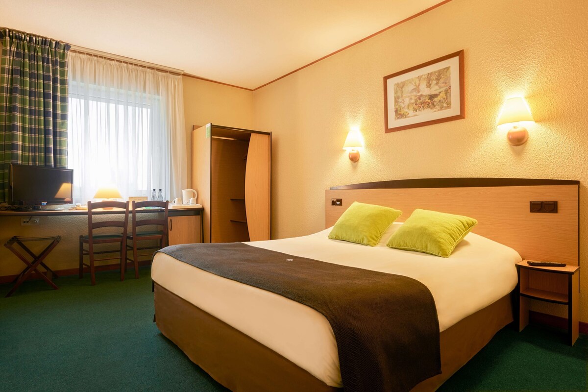 A hotel room with a bed, two chairs, a desk, a television, and two wall-mounted lamps.
