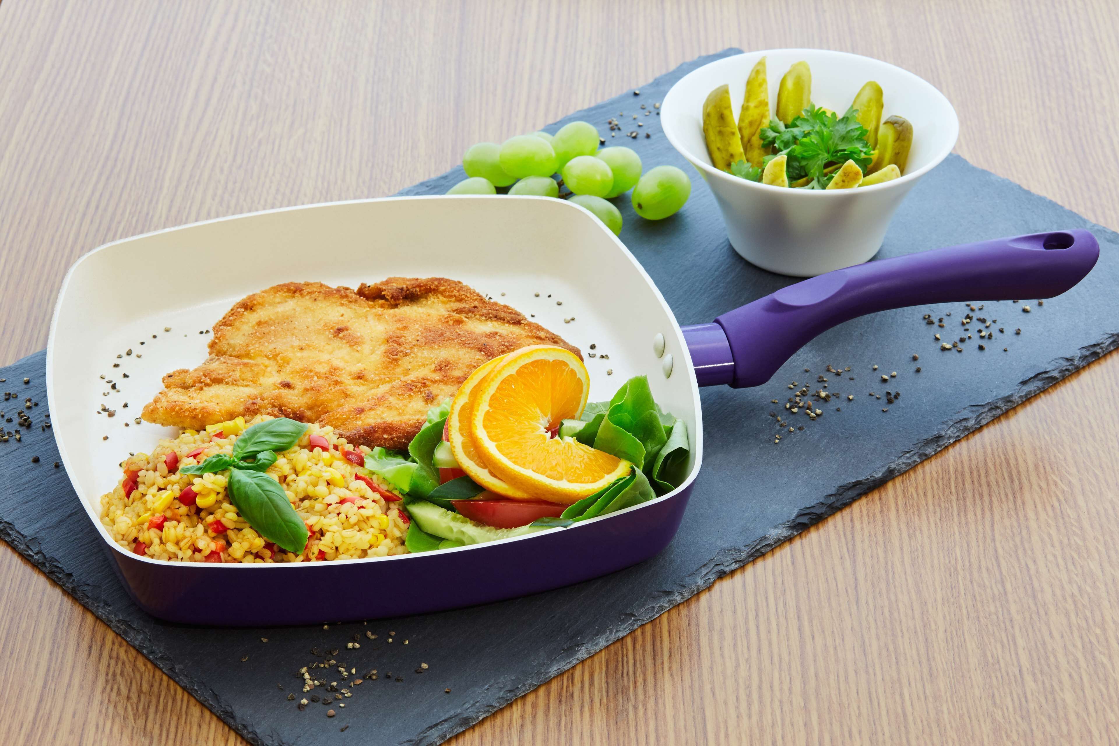 Un repas composé d'une escalope panée, de riz, de légumes, et accompagné d'une portion de cornichons et de raisins, le tout servi sur une ardoise.