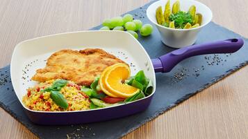 Un repas composé d'une escalope panée, de riz, de légumes, et accompagné d'une portion de cornichons et de raisins, le tout servi sur une ardoise.