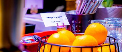 Un panier rempli d’oranges fraîches, avec une pancarte indiquant « Oranges à presser », et un récipient en métal contenant des ustensiles.