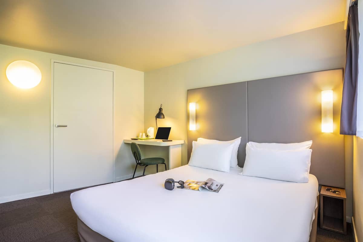 Ein Hotelzimmer mit einem Bett, einem Schreibtisch mit Laptop, einem Sessel und einem Nachttisch mit Gläsern.