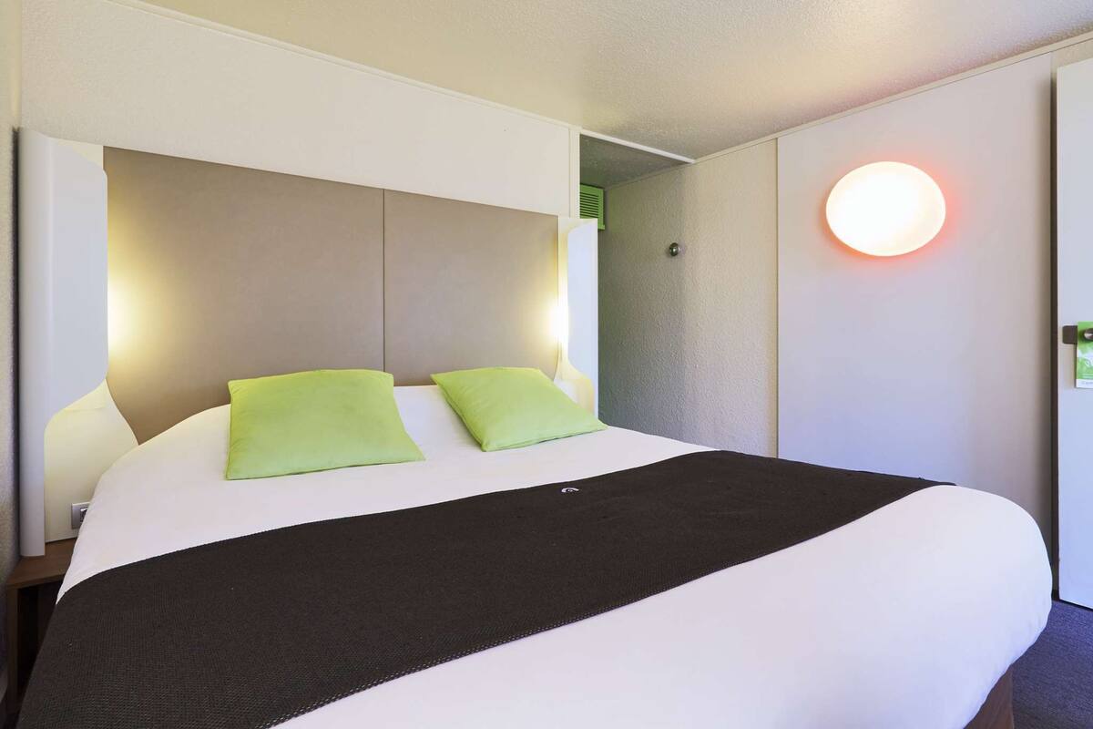 Habitación de hotel con una cama grande, dos almohadas verdes, un edredón negro y una lámpara de pared redonda.
