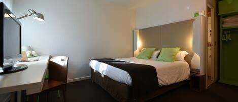 A hotel room with a bed, a desk with a chair, a lamp, and a green wardrobe.
