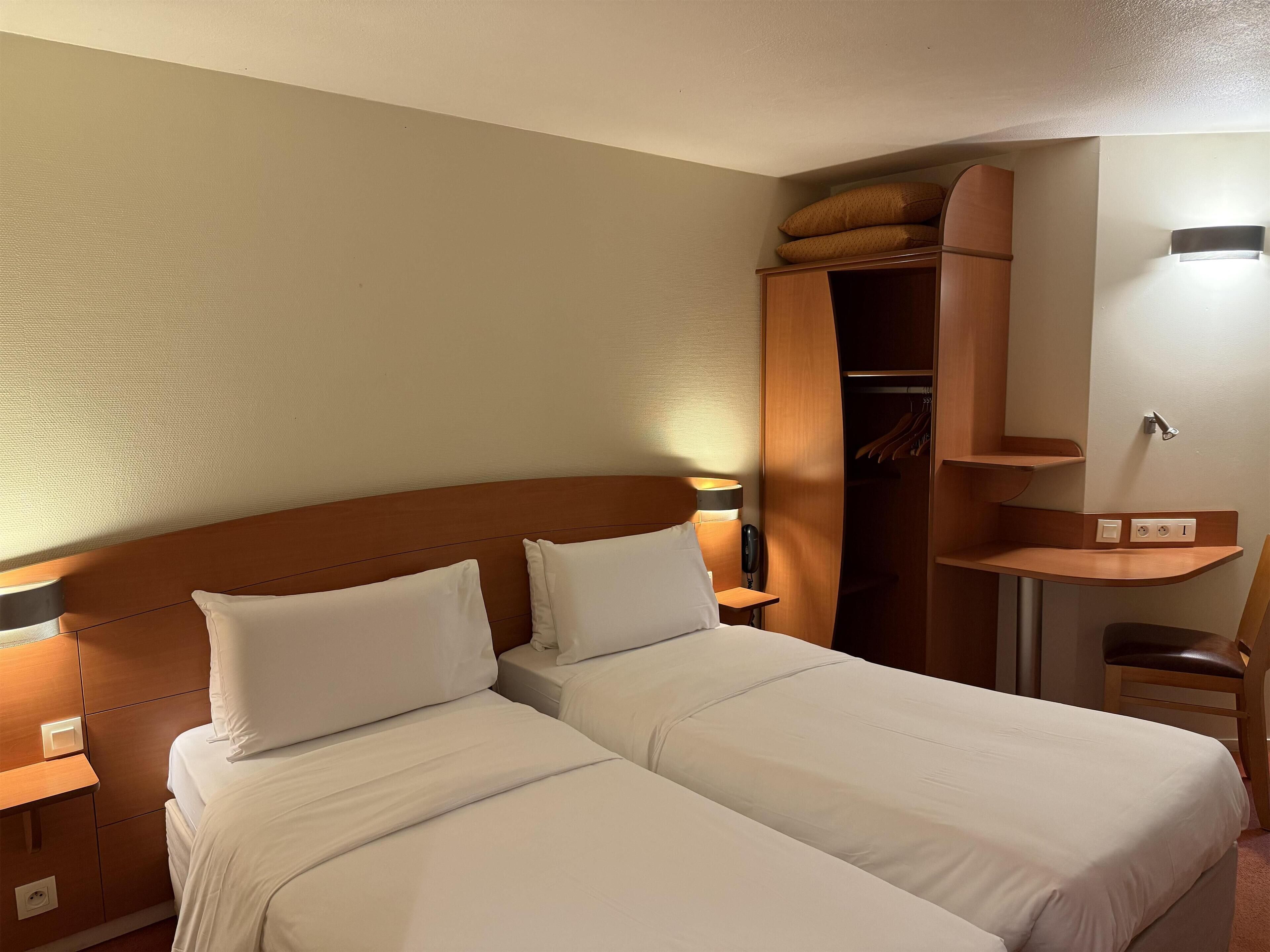 Hotellihuone, jossa on kaksi sänkyä, puinen pääty, vaatekaappi, yöpöytä lampun kanssa ja pieni hylly.