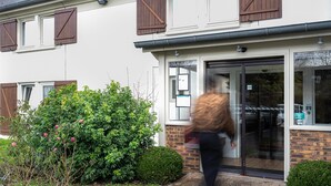 A building with a brick facade, large glass entrance doors, and a small garden with pink flowers. - Hôtel Kyriad Rennes Nord (Rennes)