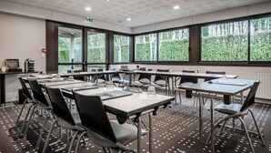 A conference room with multiple tables and chairs, large windows with a view of greenery, and a ceiling with a grid pattern. - Hôtel Kyriad Rennes Nord (Rennes)