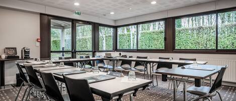 Uma sala de conferência com várias mesas e cadeiras, janelas amplas com vista para área verde e teto com padrão de grade.