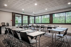 Une salle de conférence dotée de plusieurs tables et chaises, de grandes fenêtres offrant une vue sur la verdure et d’un plafond orné d’un motif en réseau.