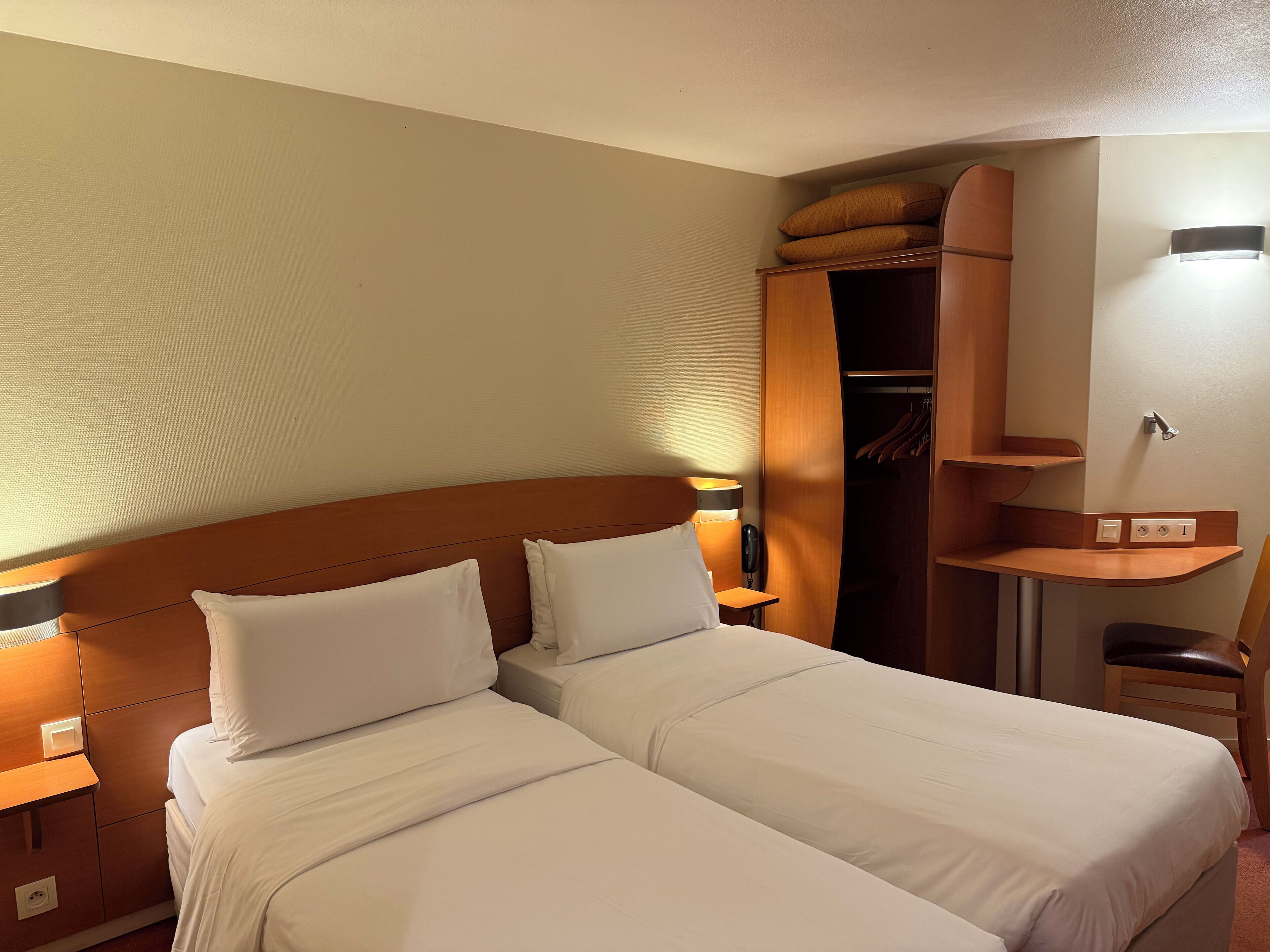 A hotel room with two beds, a wooden headboard, a wardrobe, a bedside table with a lamp, and a small desk with a chair.