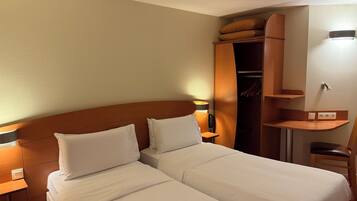 A hotel room with two beds, a wooden headboard, a wardrobe, a bedside table with a lamp, and a small desk with a chair.