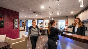 A modern hotel lobby with a reception desk, seating area, and a red brick accent wall. - Hôtel Kyriad Rennes Nord (Rennes)