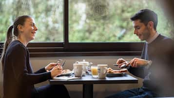 Una mesa de comedor con alimentos para el desayuno, una ventana con vistas a la vegetación, y una persona usando un teléfono inteligente.