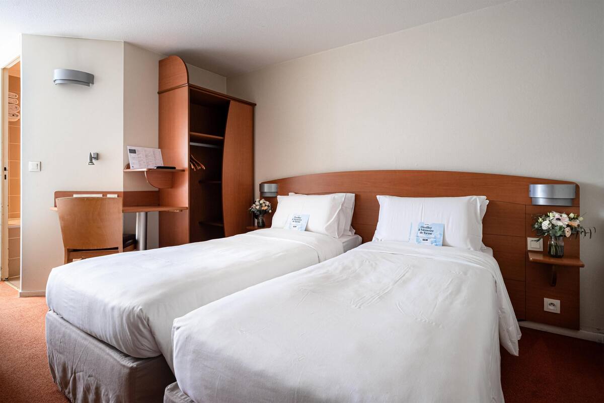 A hotel room with two single beds, a wooden headboard, a small desk, and a wardrobe.