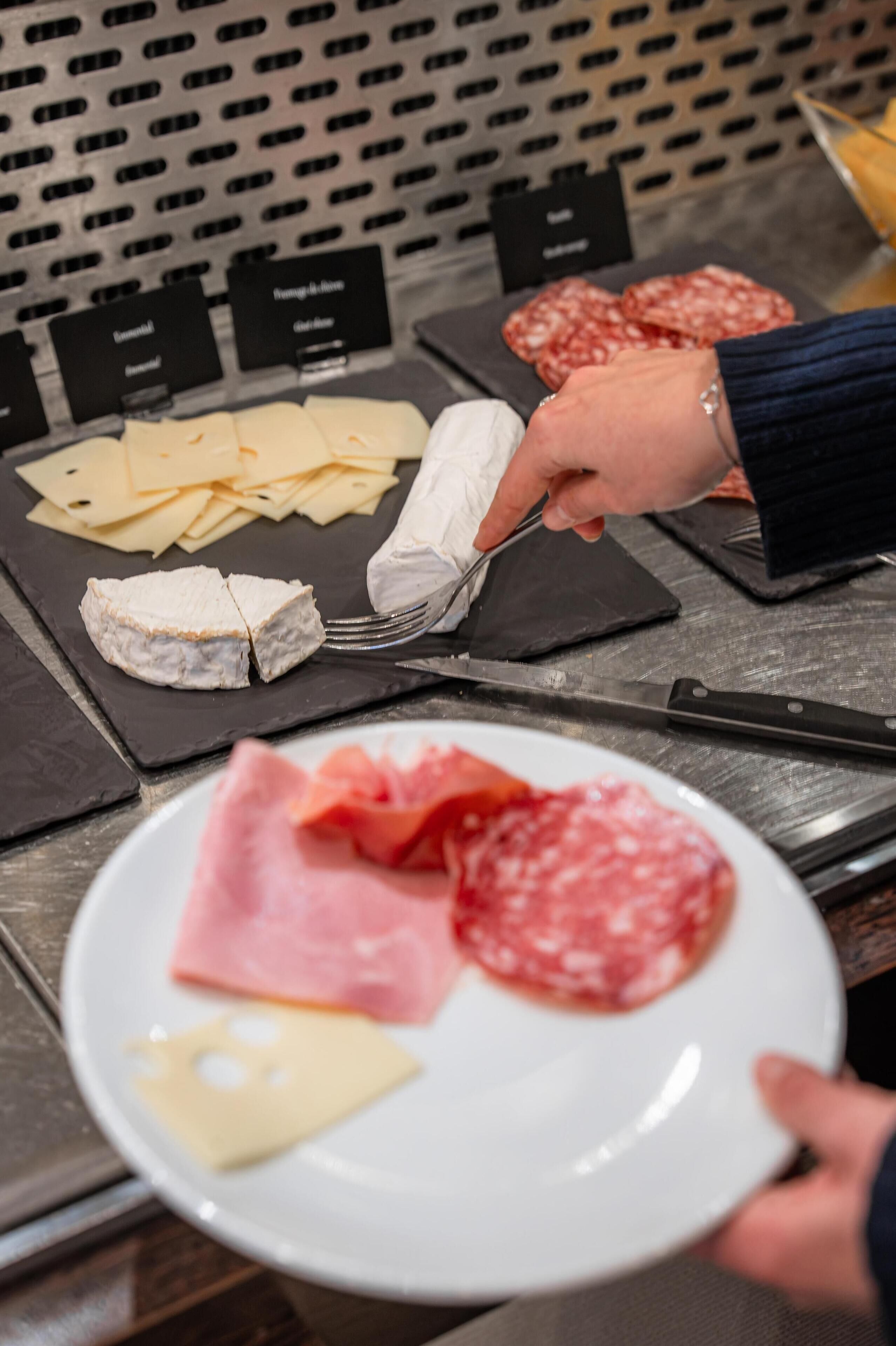 A person holding a plate with various meats and cheese, with a knife and fork on a black slate board in the background.