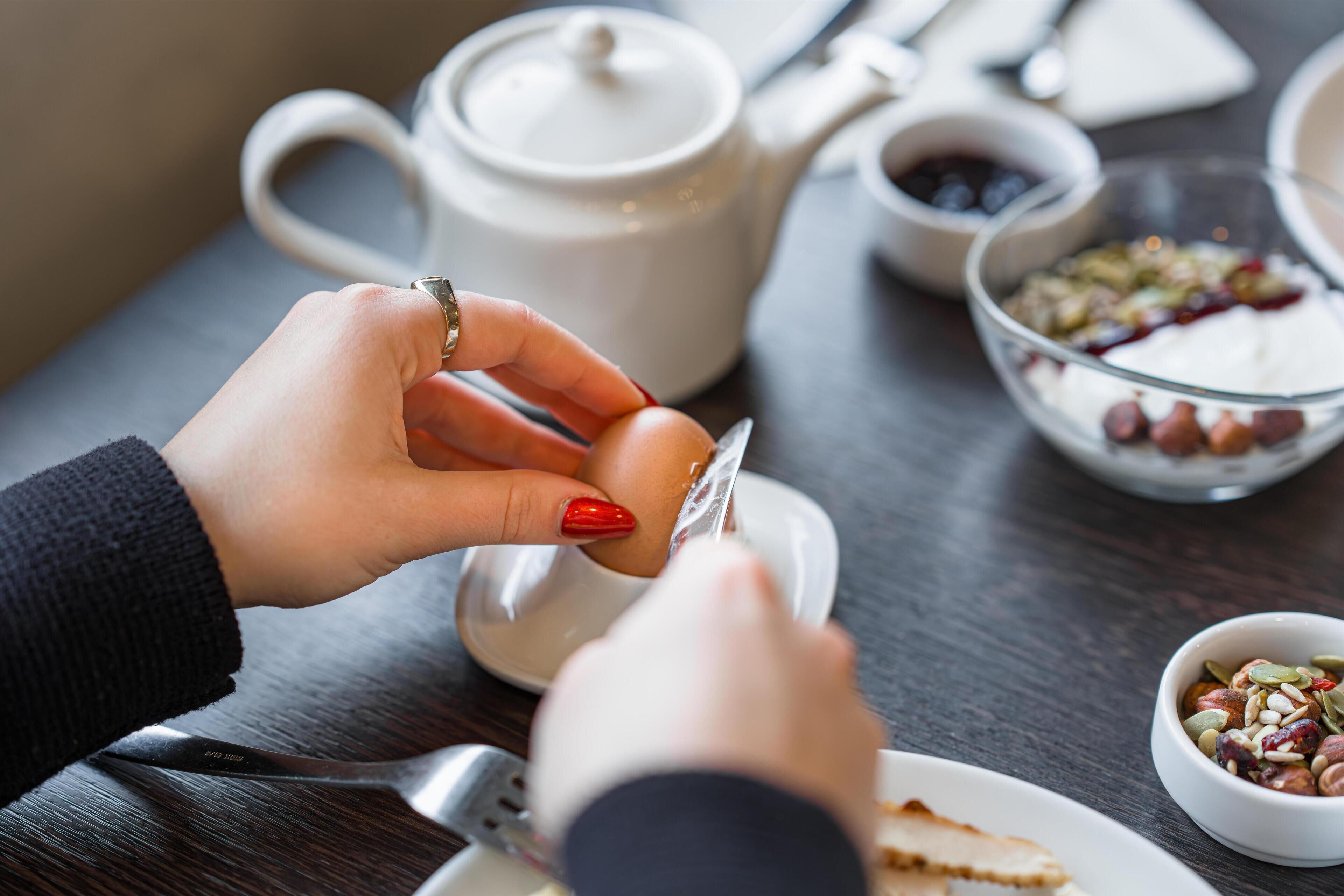 A person peeling an egg over a white plate, with a teapot and bowls of food in the background.
