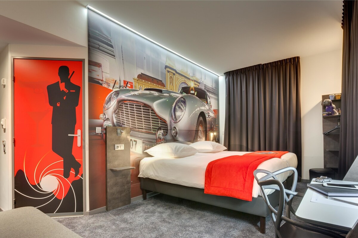 Una habitación de hotel moderna con una cama, un escritorio y una pared decorada con un coche antiguo y la silueta de una persona.
