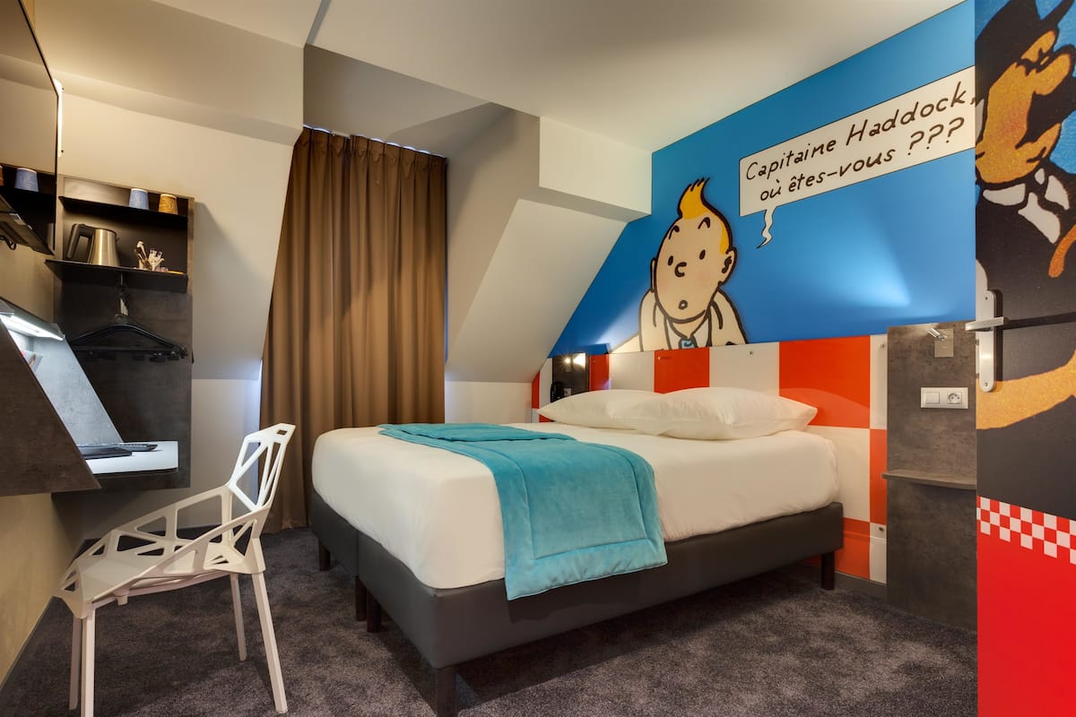 A hotel room with a bed, a desk, a chair, and a wall with a cartoon character.