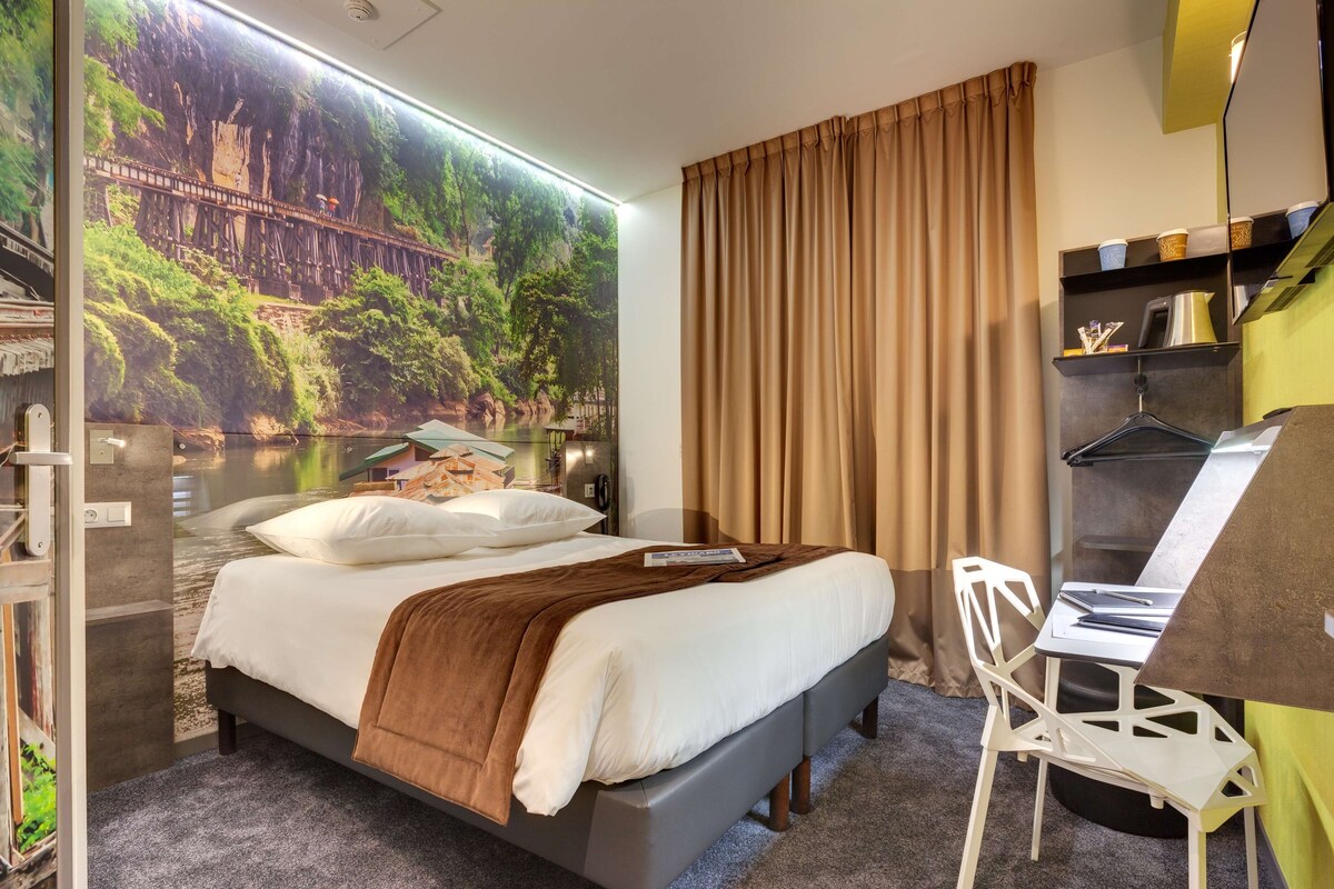 Habitación de hotel con cama, escritorio y un amplio ventanal con vistas panorámicas a un puente y un bosque.