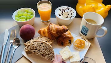 Una mesa de desayuno con cruasanes, queso, jamón, pan, uvas, zumo de naranja, café y un juguete con forma de pato.