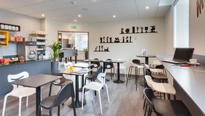 Una cafetería moderna con mesas y sillas, una barra con un ordenador portátil, y una pared con varios objetos decorativos.