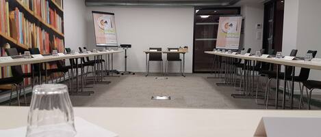Un salón de conferencias con mesas largas, sillas, una pantalla de proyector y una estantería.