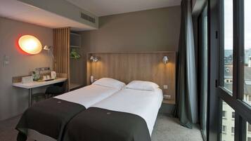 A hotel room with a bed, a desk, a chair, a lamp, and a view of the city through the window.