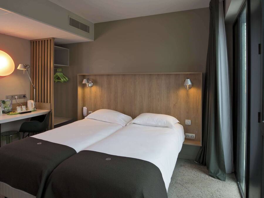 A hotel room with a bed, a desk, a chair, a lamp, and a view of the city through the window.