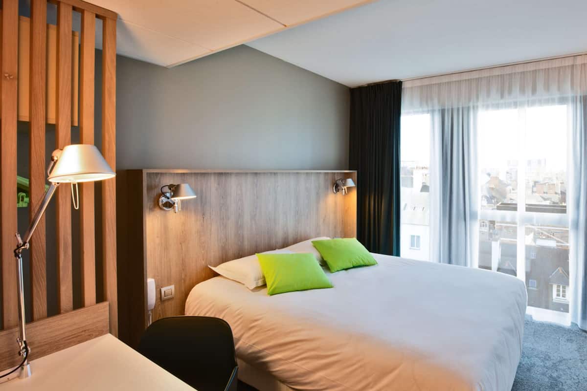 A hotel room with a large bed, two bedside lamps, a wooden headboard, a desk with a chair, and a view of the city through large windows.