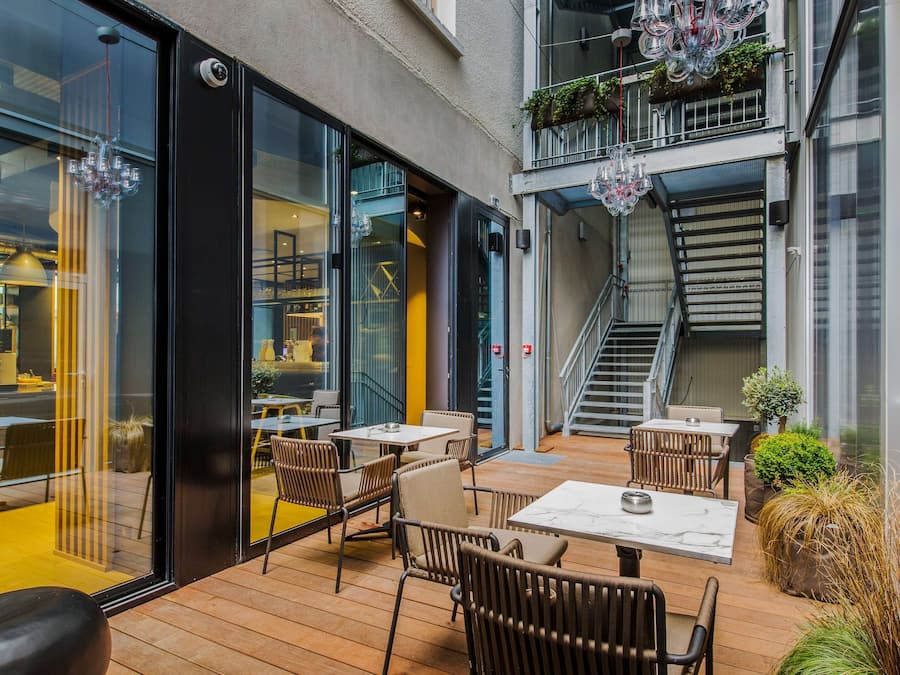 An outdoor patio with wooden flooring, tables, and chairs. There are glass doors leading to an interior space with a yellow wall and a staircase.