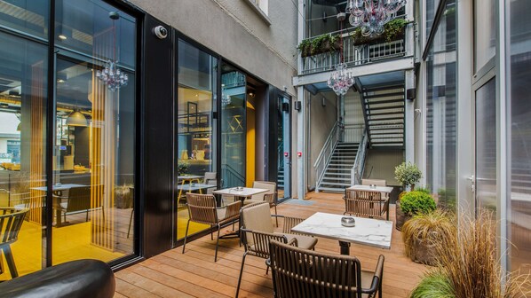 Un patio exterior con suelo de madera, mesas y sillas. Hay puertas de cristal que dan a un espacio interior con una pared amarilla y una escalera.
