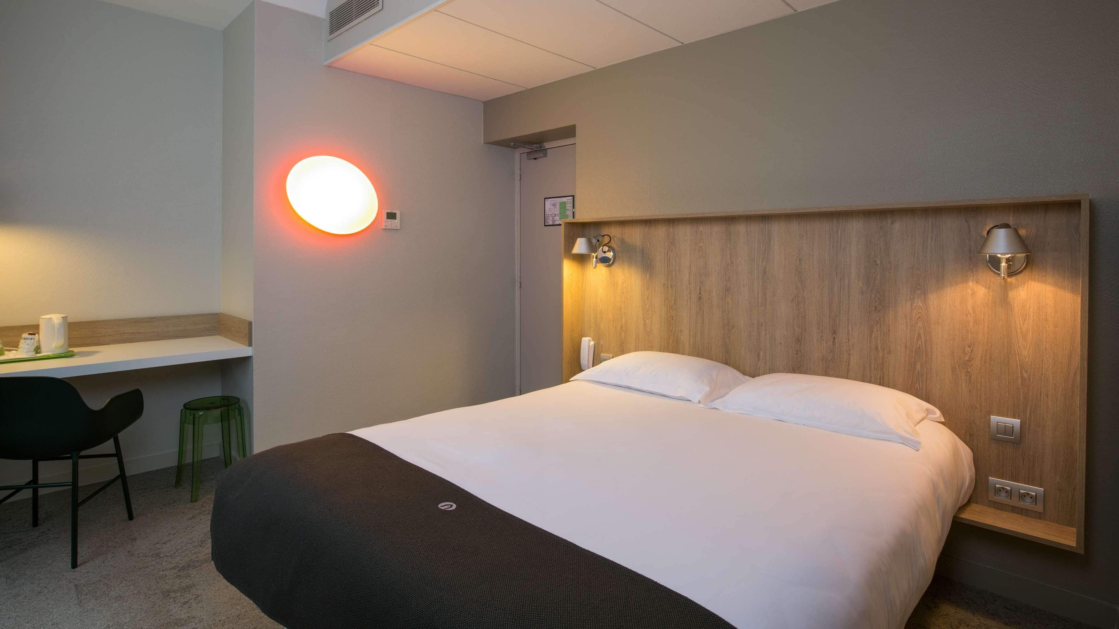 A hotel room with a large bed, a desk with a chair, and a wall-mounted lamp.
