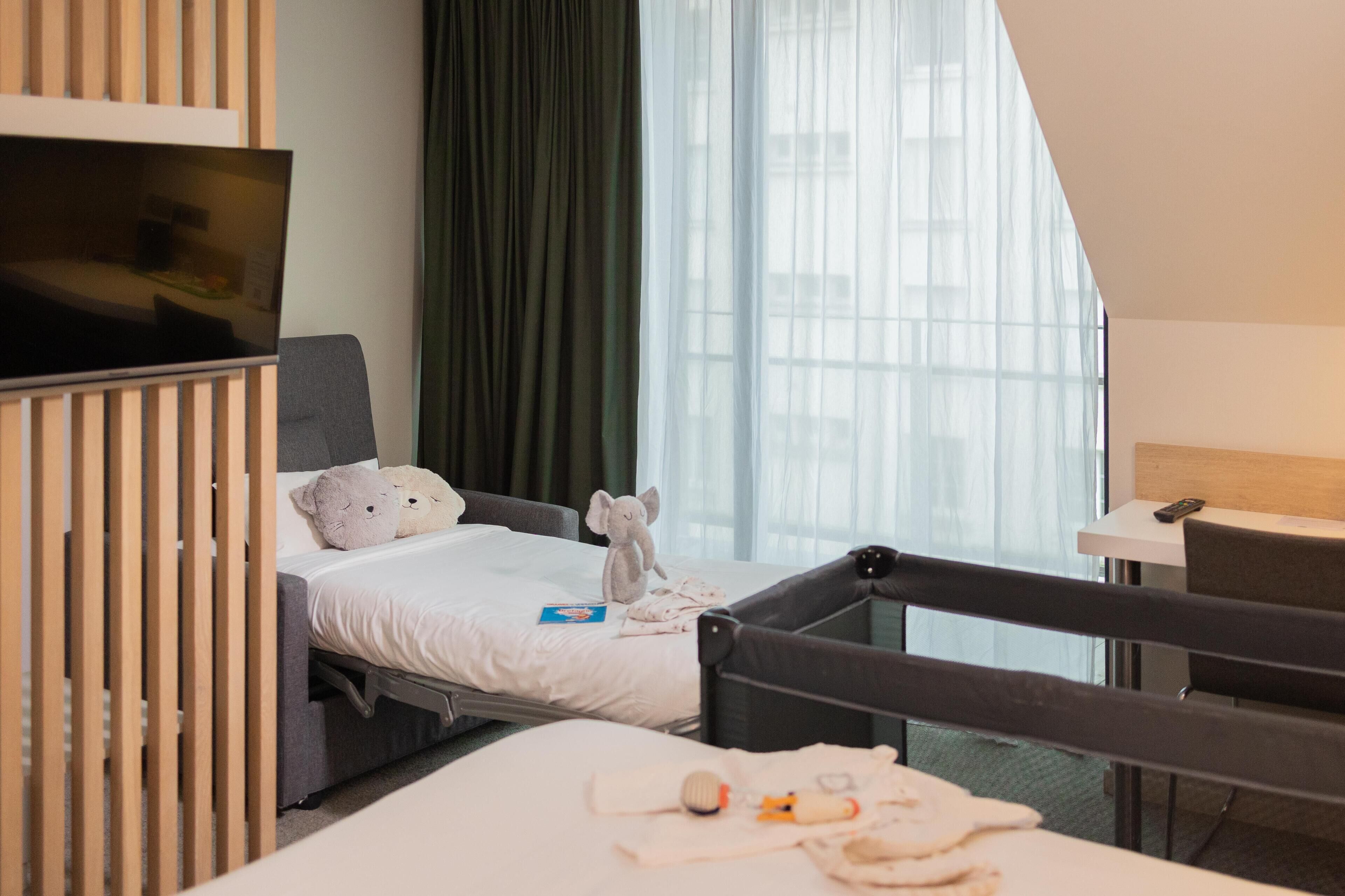 Una habitación de hotel con cama, una silla, televisor y ventana con cortinas.