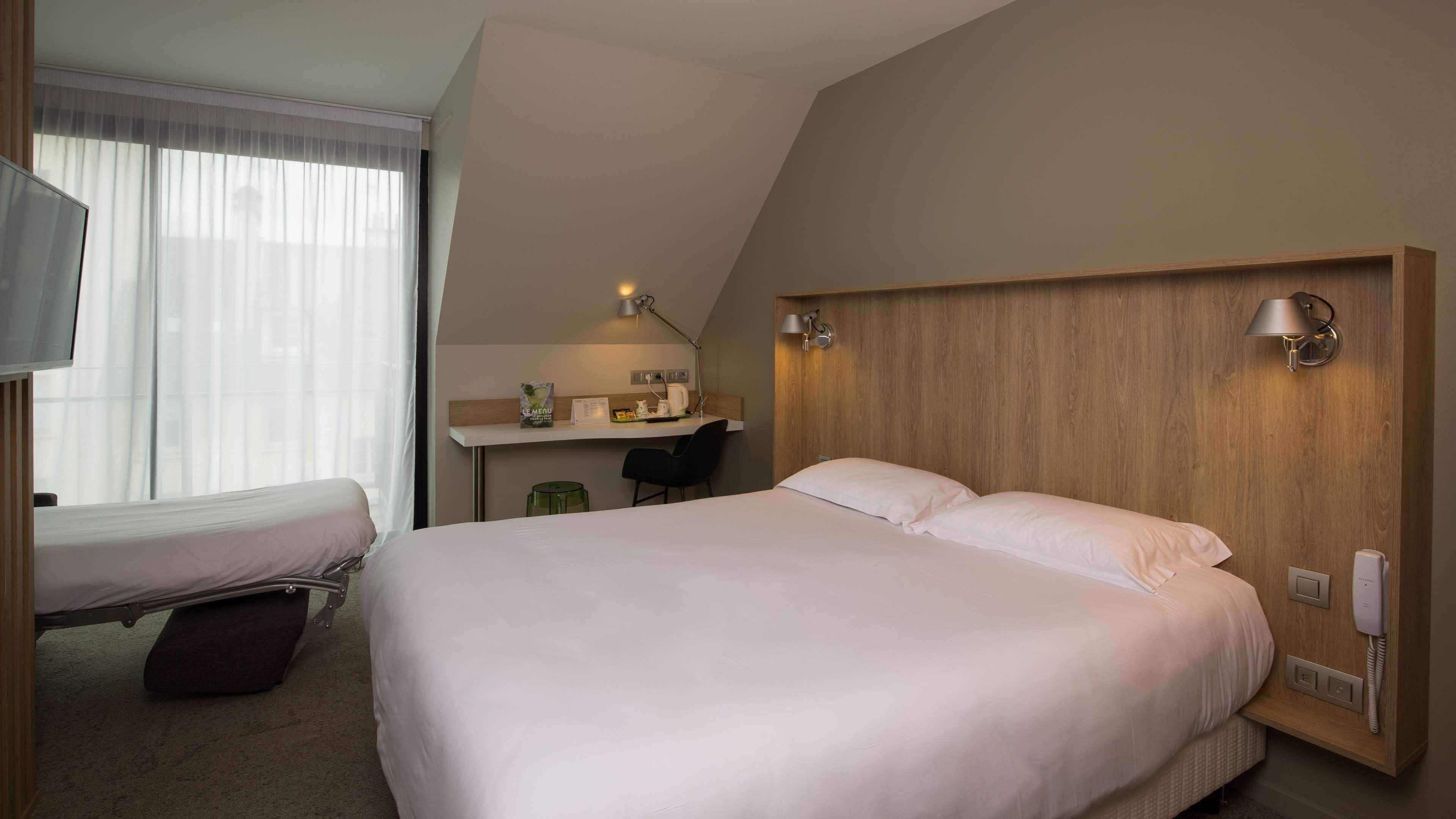 Habitación de hotel con una cama grande, una cama más pequeña, un escritorio con silla, un televisor y una ventana con cortinas translúcidas.
