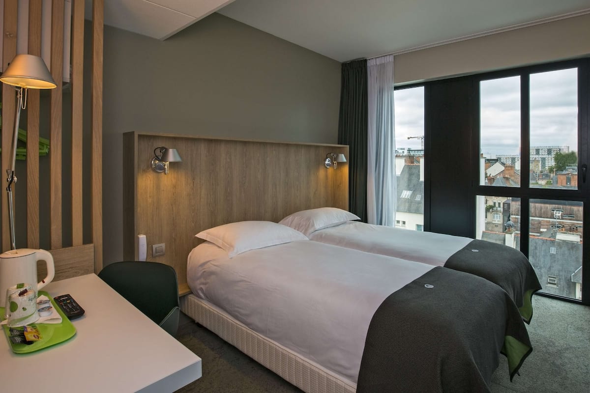 A hotel room with two beds, a desk with a lamp, a coffee maker, and a view of the city through large windows.